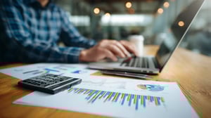 Empresário analisando relatórios financeiros e gráficos no laptop, calculadora e documentos sobre mesa de madeira, escritório moderno ao fundo, foco em gráficos de crescimento de receita.